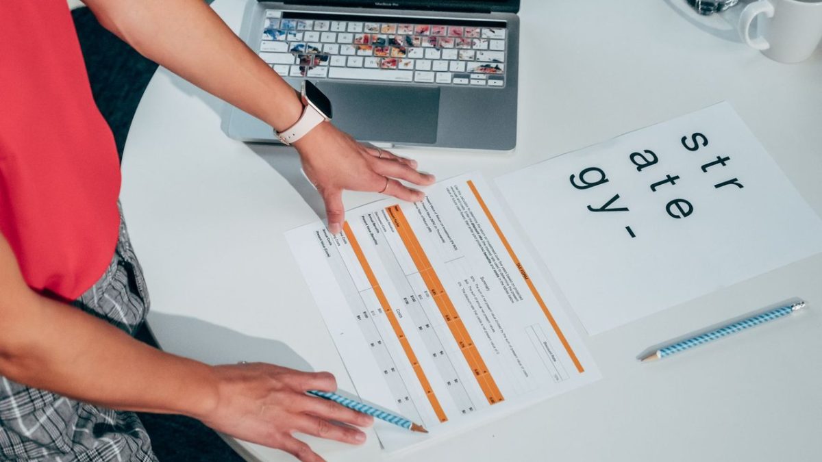 Pessoa analisando documentos e um laptop sobre uma mesa, em uma atividade de planejamento e avaliação de projetos.