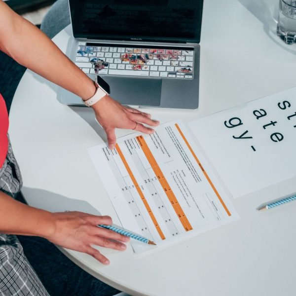 Pessoa analisando documentos e um laptop sobre uma mesa, em uma atividade de planejamento e avaliação de projetos.