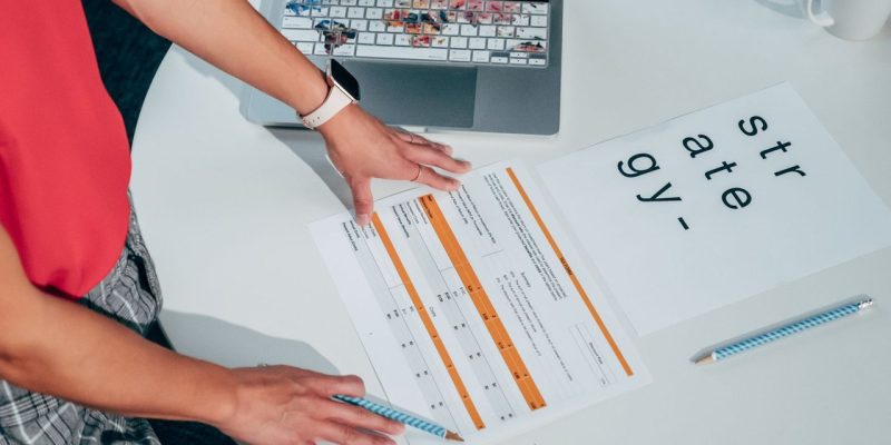 Pessoa analisando documentos e um laptop sobre uma mesa, em uma atividade de planejamento e avaliação de projetos.