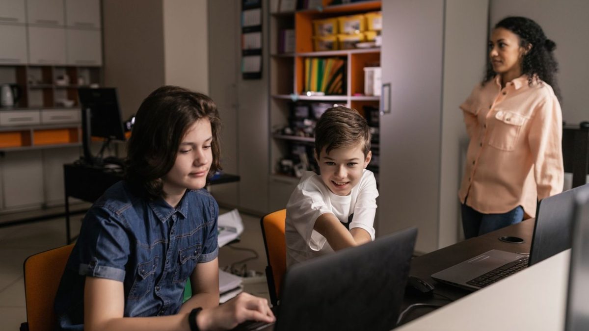 Estudantes usam laptops em sala de aula enquanto uma professora os supervisiona.