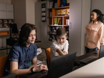 Estudantes usam laptops em sala de aula enquanto uma professora os supervisiona.