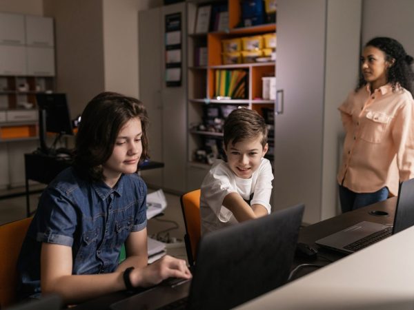 Estudantes usam laptops em sala de aula enquanto uma professora os supervisiona.