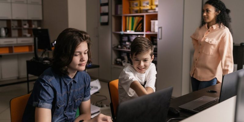 Estudantes usam laptops em sala de aula enquanto uma professora os supervisiona.