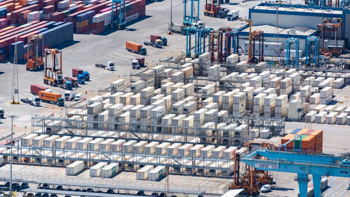 Vista aérea de um porto de cargas com contêineres empilhados e máquinas de movimentação logística.