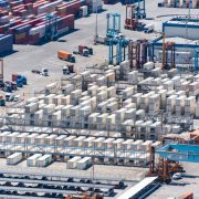 Vista aérea de um porto de cargas com contêineres empilhados e máquinas de movimentação logística.