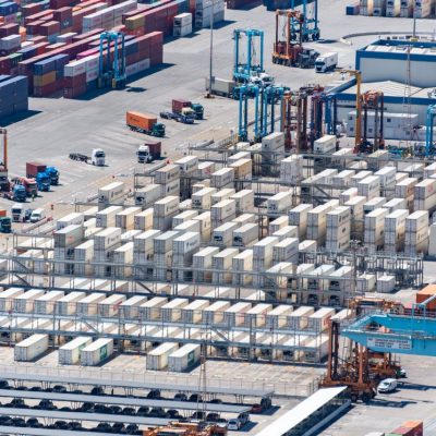 Vista aérea de um porto de cargas com contêineres empilhados e máquinas de movimentação logística.