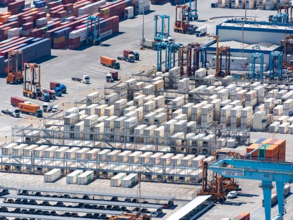 Vista aérea de um porto de cargas com contêineres empilhados e máquinas de movimentação logística.