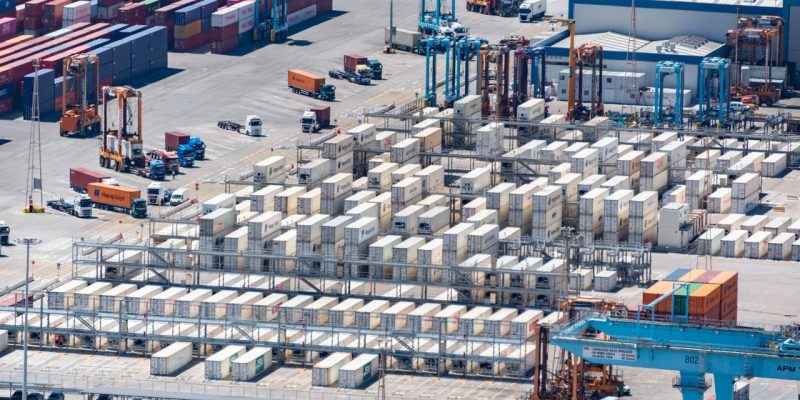 Vista aérea de um porto de cargas com contêineres empilhados e máquinas de movimentação logística.